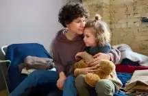 Younger foster mom giving hug to toddler foster girl holding a brown fuzzy teddybear