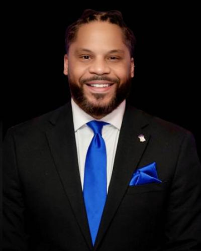 Wil Brown in a black suit with a blue tie with a black background